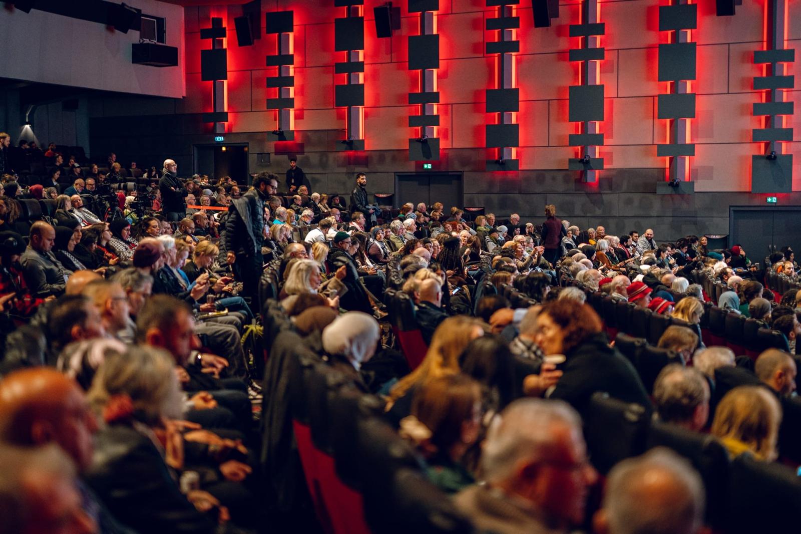 Packed conference hall seen from the side, with the audience in front of a red-lit acoustic wall.