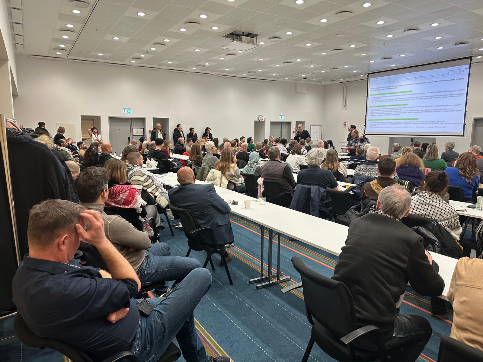 Seminar room with participants at long tables and a presentation slide on the screen.