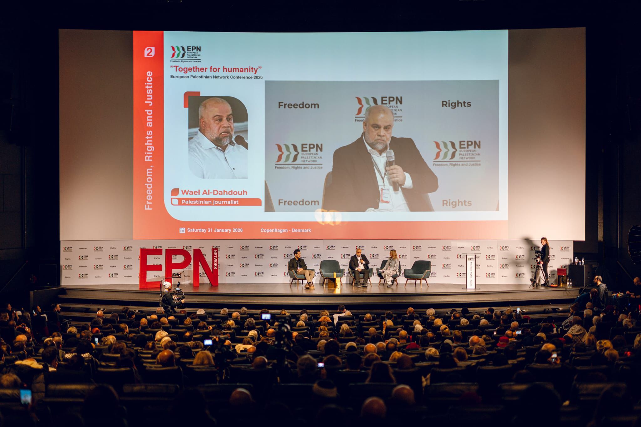 Main stage with the “Together for humanity” screen showing a portrait of Wael Al-Dahdouh; a speaker at the microphone with the panel seated behind.
